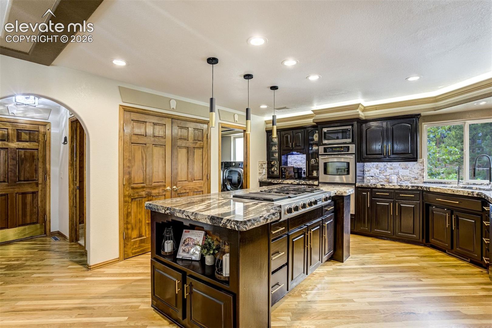Kitchen featuring a large island with a cooktop, dark wood cabinetry, integrated stainless steel appliances, stone countertops, and wood-finish flooring