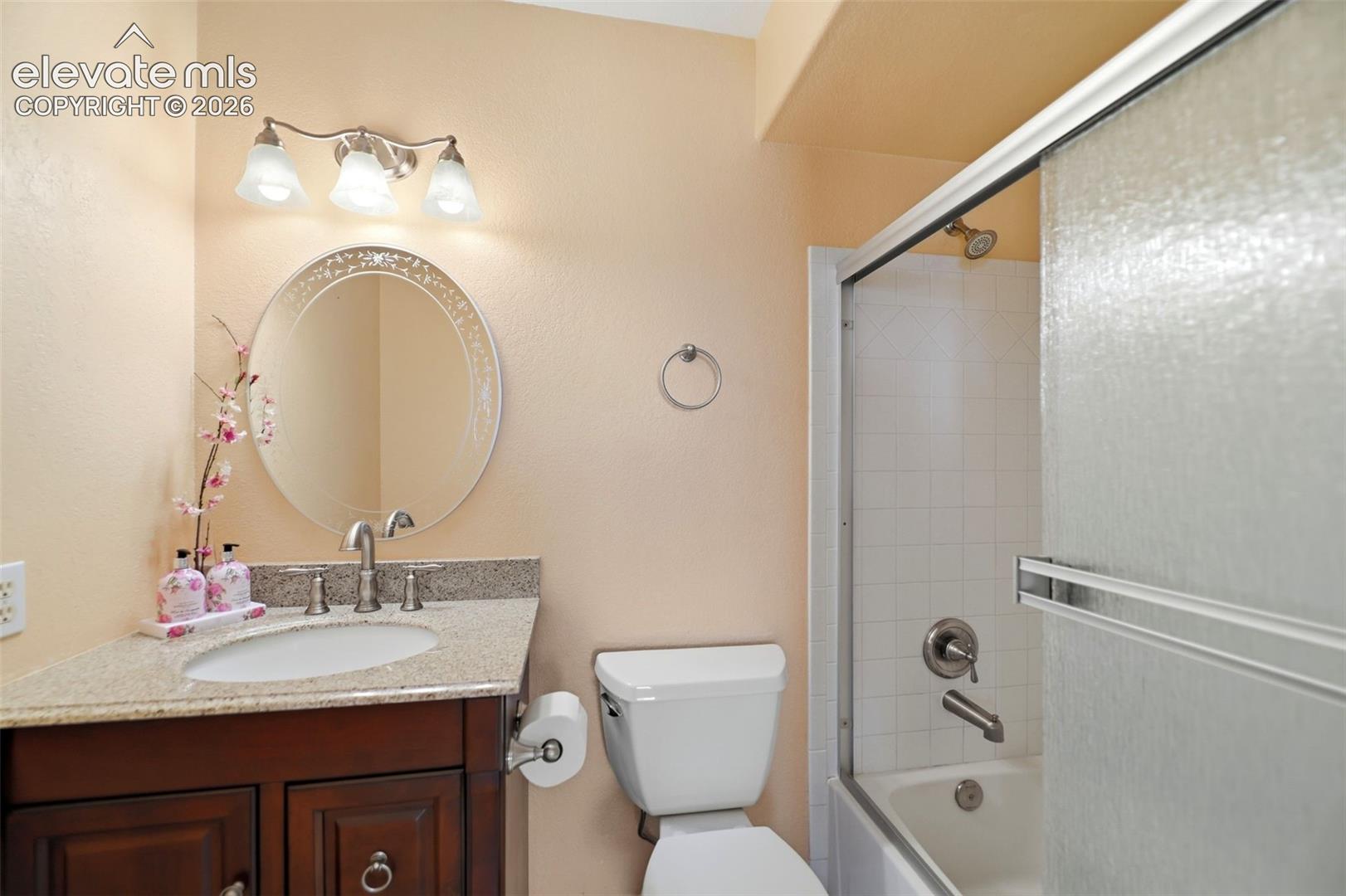 Bathroom featuring a dark wood vanity with a light speckled countertop, an oval-shaped mirror, a three-light vanity fixture, and a bathtub with white subway tile surround and frosted sliding glass doors