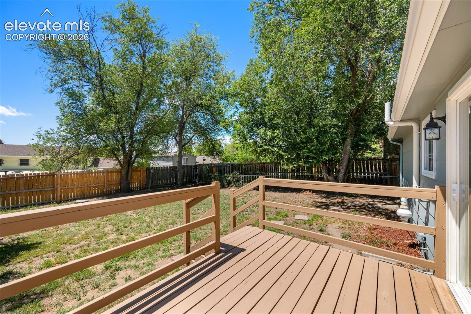 Wood-finish deck with railing, spacious yard, mature trees, wood privacy fence, and exterior wall sconce