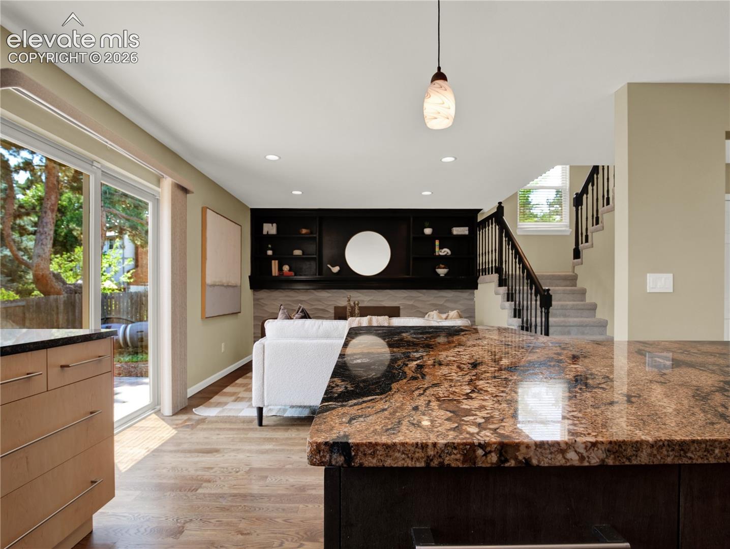 Polished stone countertop with waterfall edge, built-in black cabinetry with display shelving, sliding glass door, recessed lighting, and wooden staircase with decorative metal railing
