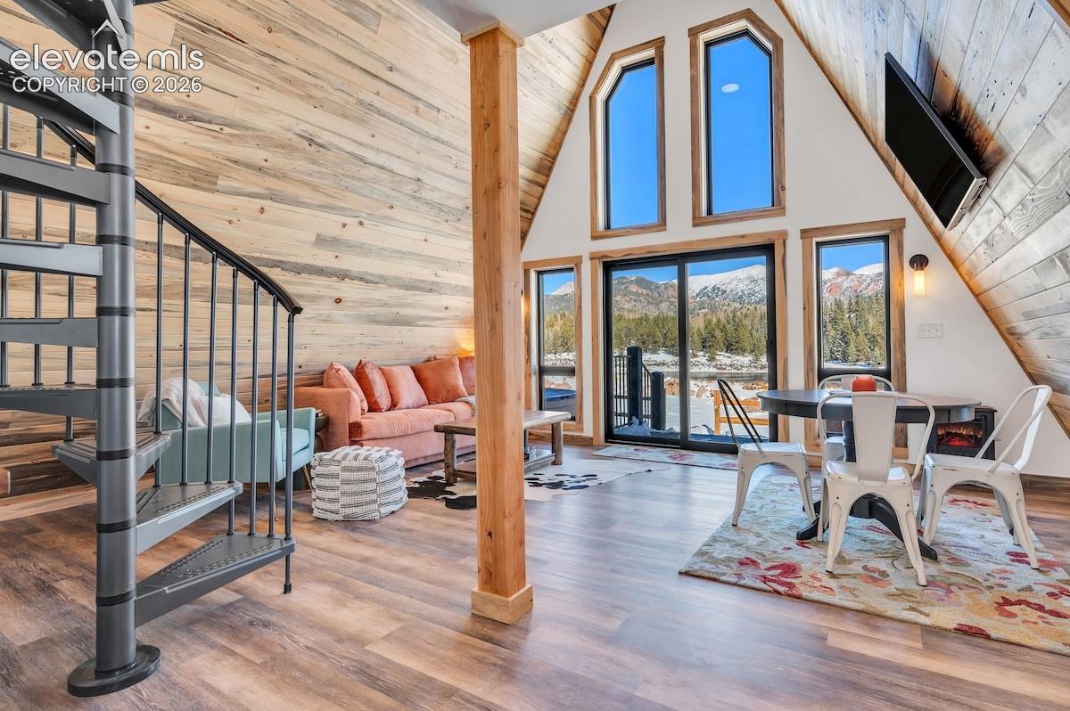 A-frame interior featuring natural wood-paneled vaulted ceilings, full-height windows, a metal spiral staircase, and mountain scenery