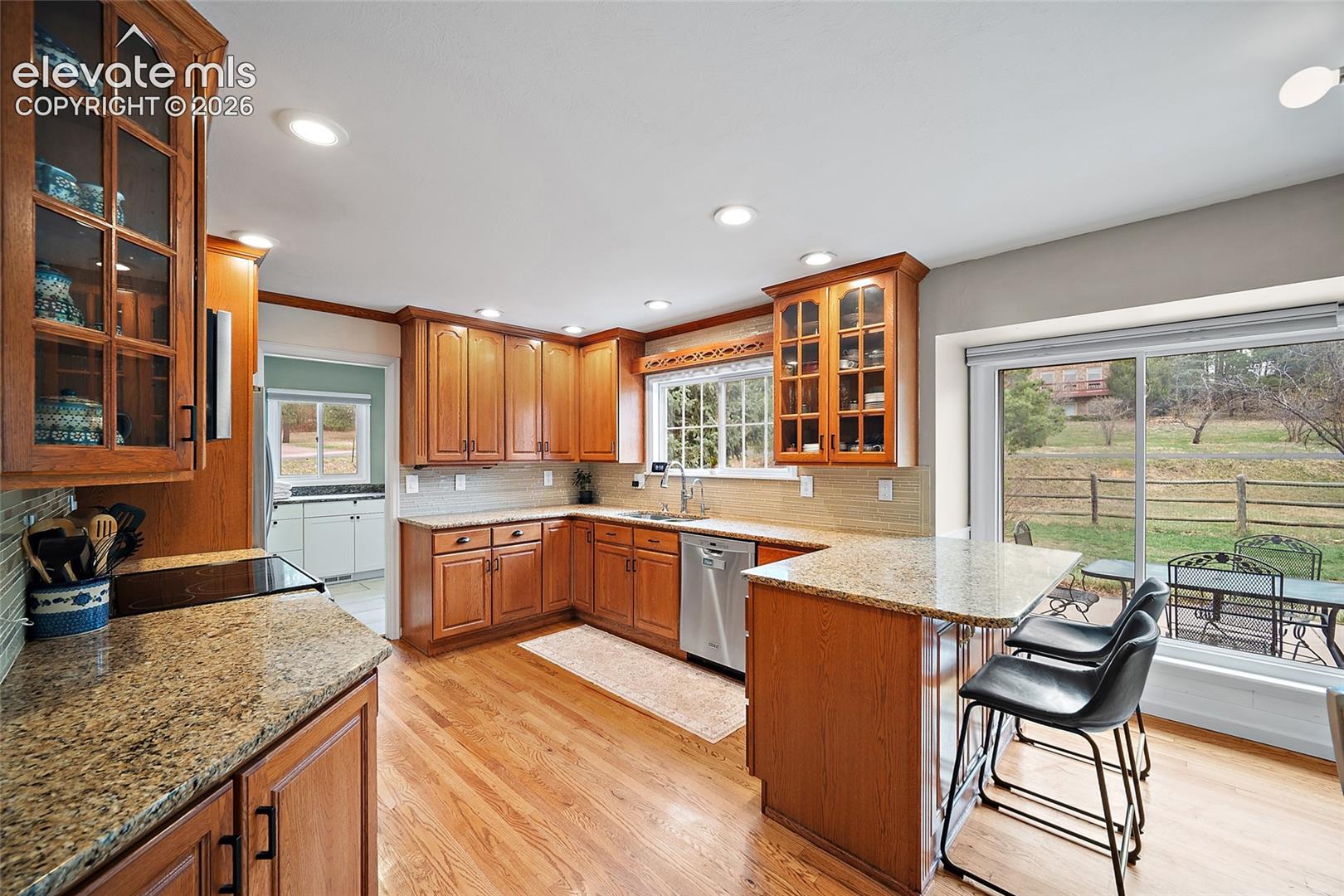 Kitchen featuring wood cabinetry with glass-front display uppers, stone countertops, stainless steel dishwasher, wood-finish flooring, and a large sliding glass door
