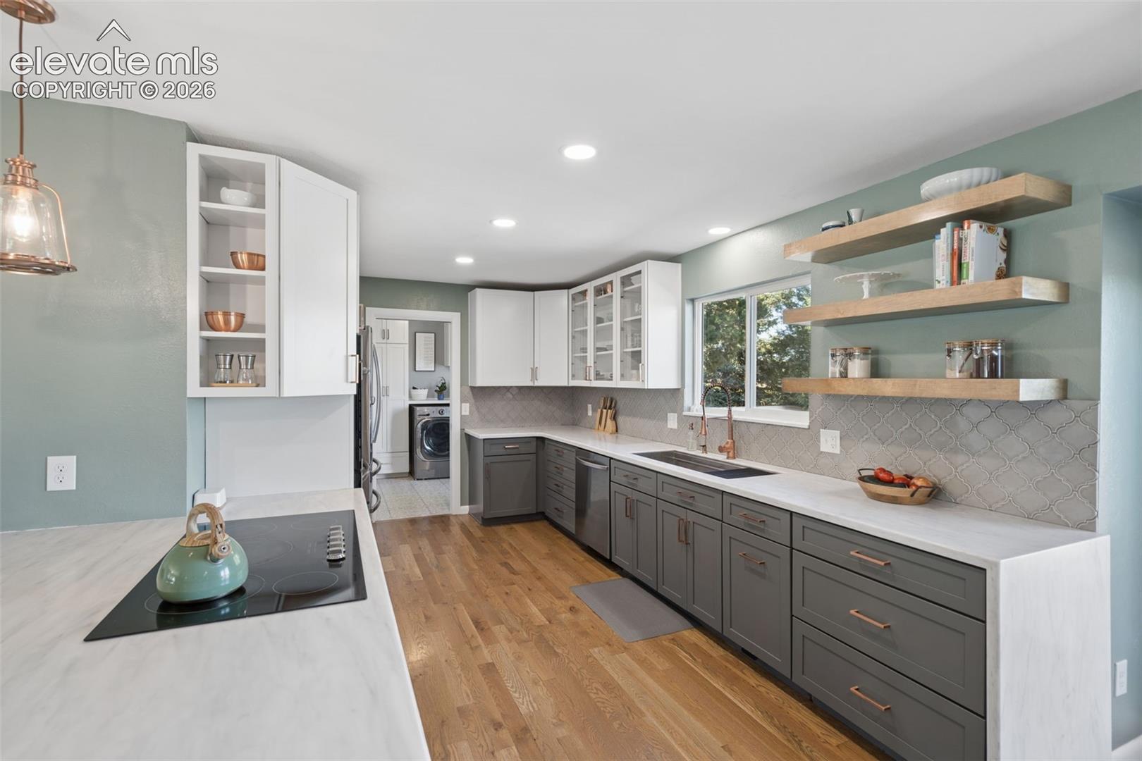 Kitchen featuring wood-finish flooring, two-tone cabinetry with contrasting white and charcoal finishes, light-toned countertops, decorative tile backsplash, and natural wood floating shelves