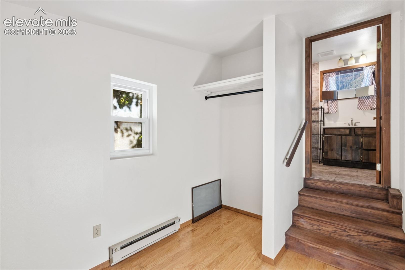 Room featuring wood-finish flooring, a built-in closet with overhead shelving, a single-pane window, and wooden steps with a metal handrail