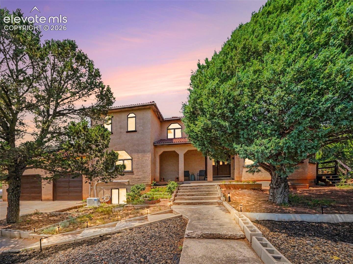 The stucco front exterior is framed by mature trees and natural landscaping, creating a welcoming entrance