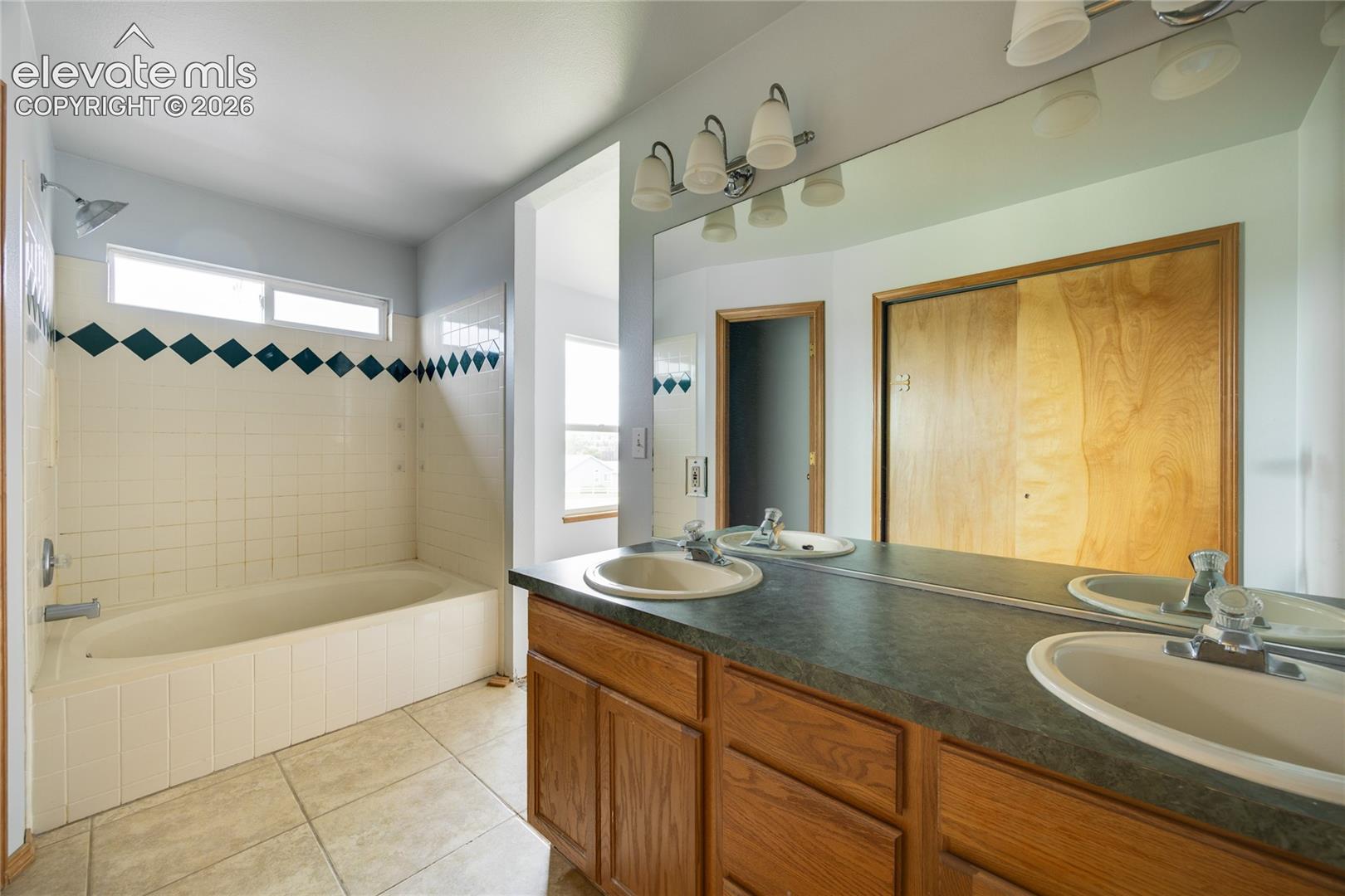 Dual vanity with integrated sinks, wood-finish cabinetry, full-width mirror, tile flooring, and a shower-tub combination with tile surround