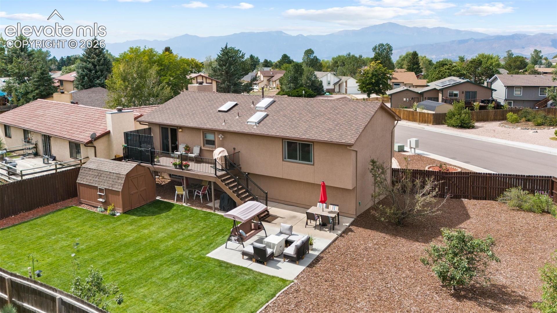 Exterior rear elevation featuring a raised deck with metal railings, a ground-level concrete patio, a detached shed, a fenced backyard with lawn, and a mountain backdrop
