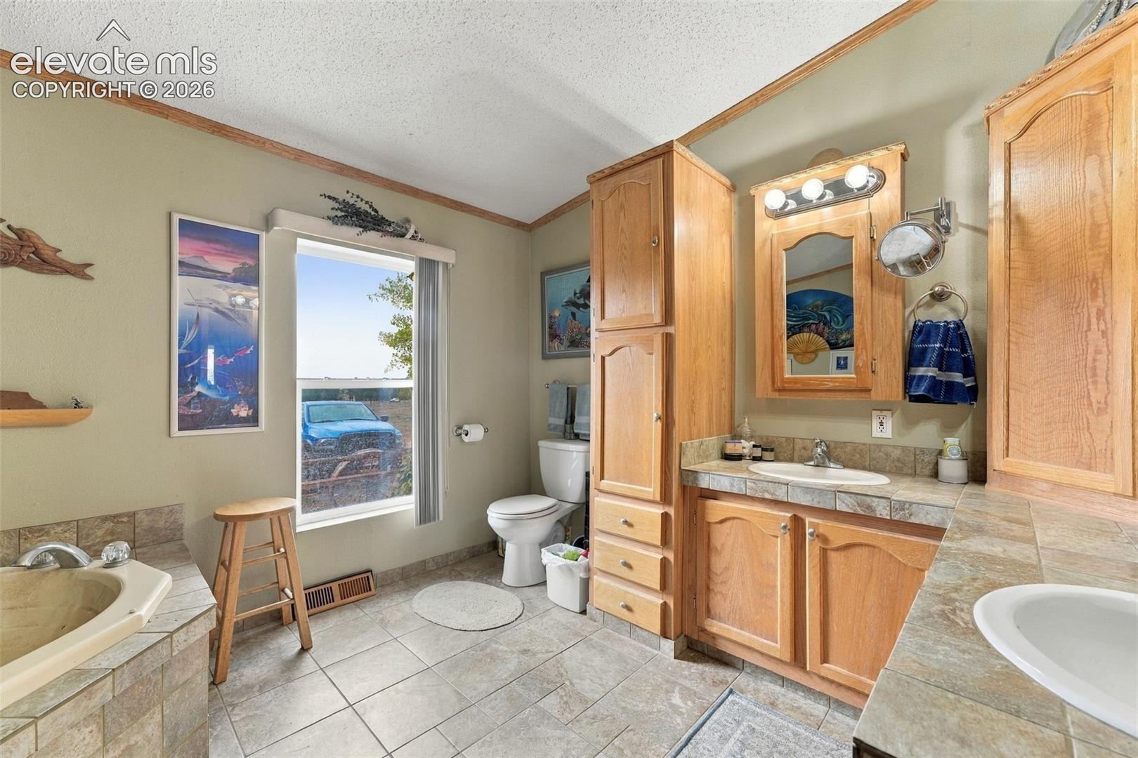 Bathroom featuring dual-sink vanity with wood cabinetry and tile countertops, tile flooring, built-in floor-to-ceiling storage, and a tile-surround soaking tub