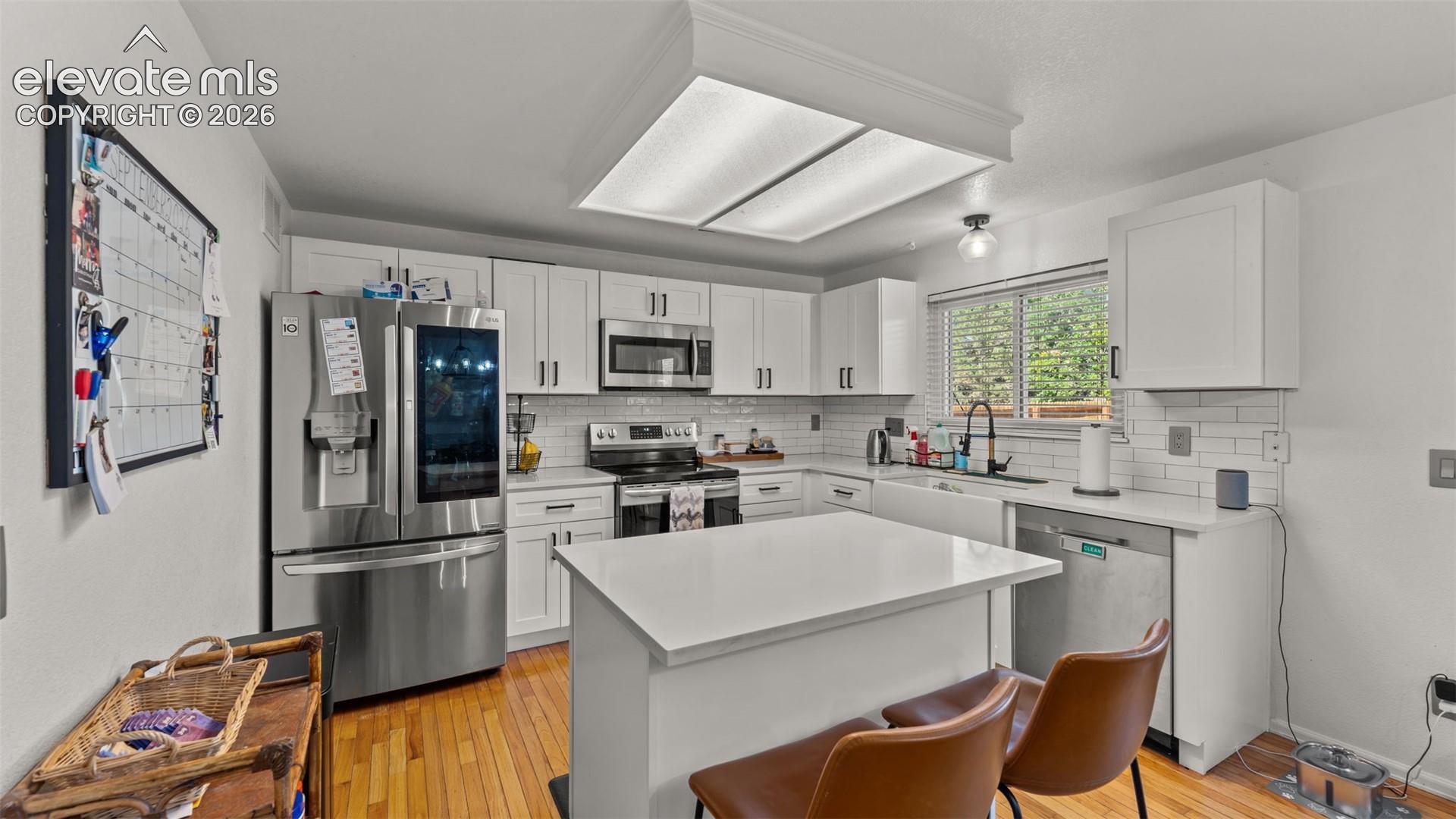 White shaker cabinetry, stainless steel appliances, central island with seating, farmhouse sink, and wood-finish flooring
