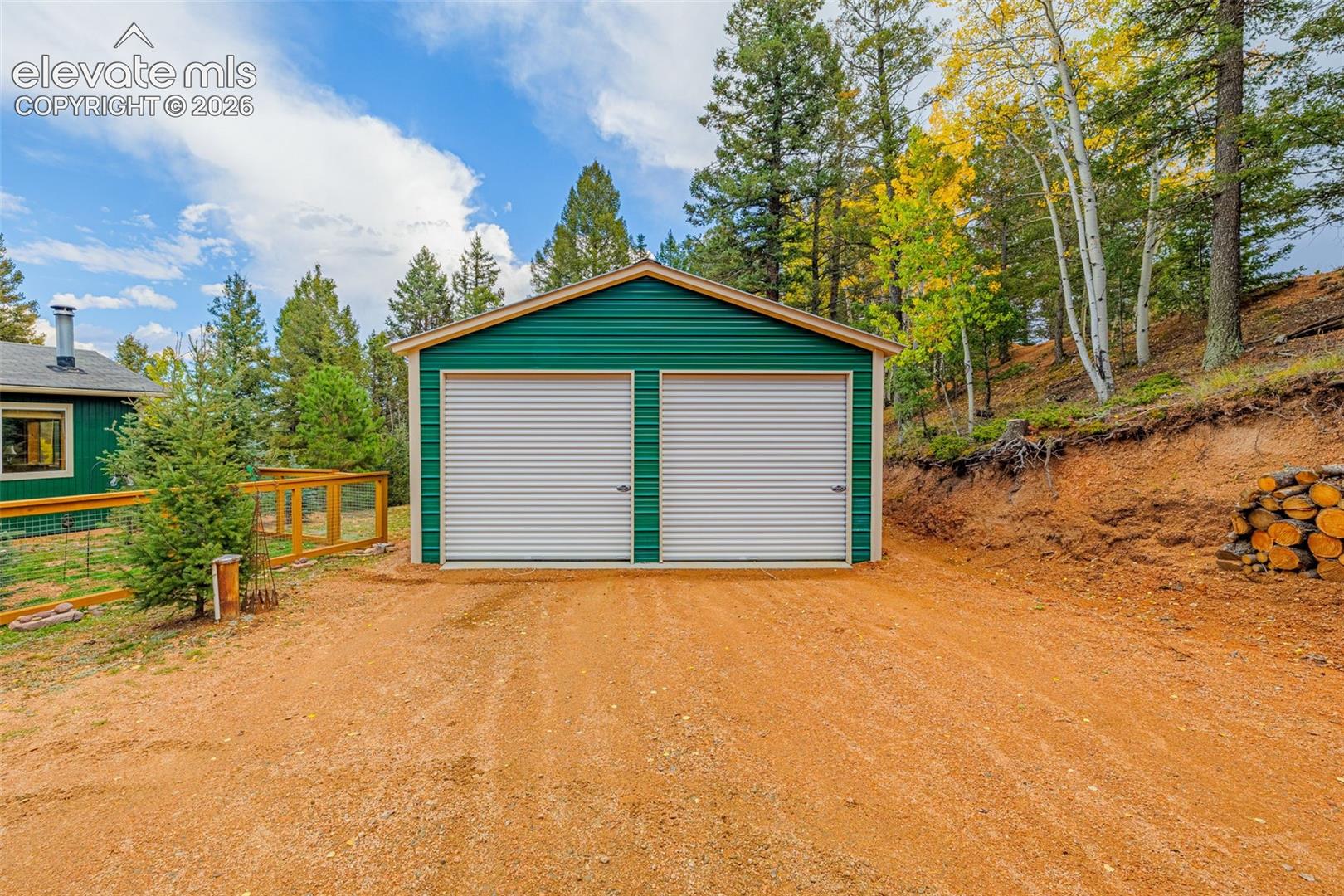 Detached two-car garage with metal siding, dual roll-up doors, and a gable roofline situated on a graded lot with natural terrain and perimeter wood fencing