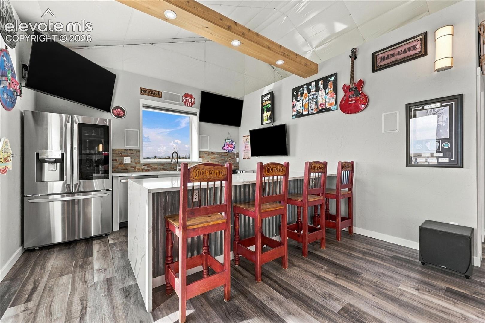 Custom bar area featuring wood-finish flooring, a stainless steel refrigerator, a corrugated metal base island with a countertop, stone tile backsplash, and a vaulted ceiling with a structural timber beam