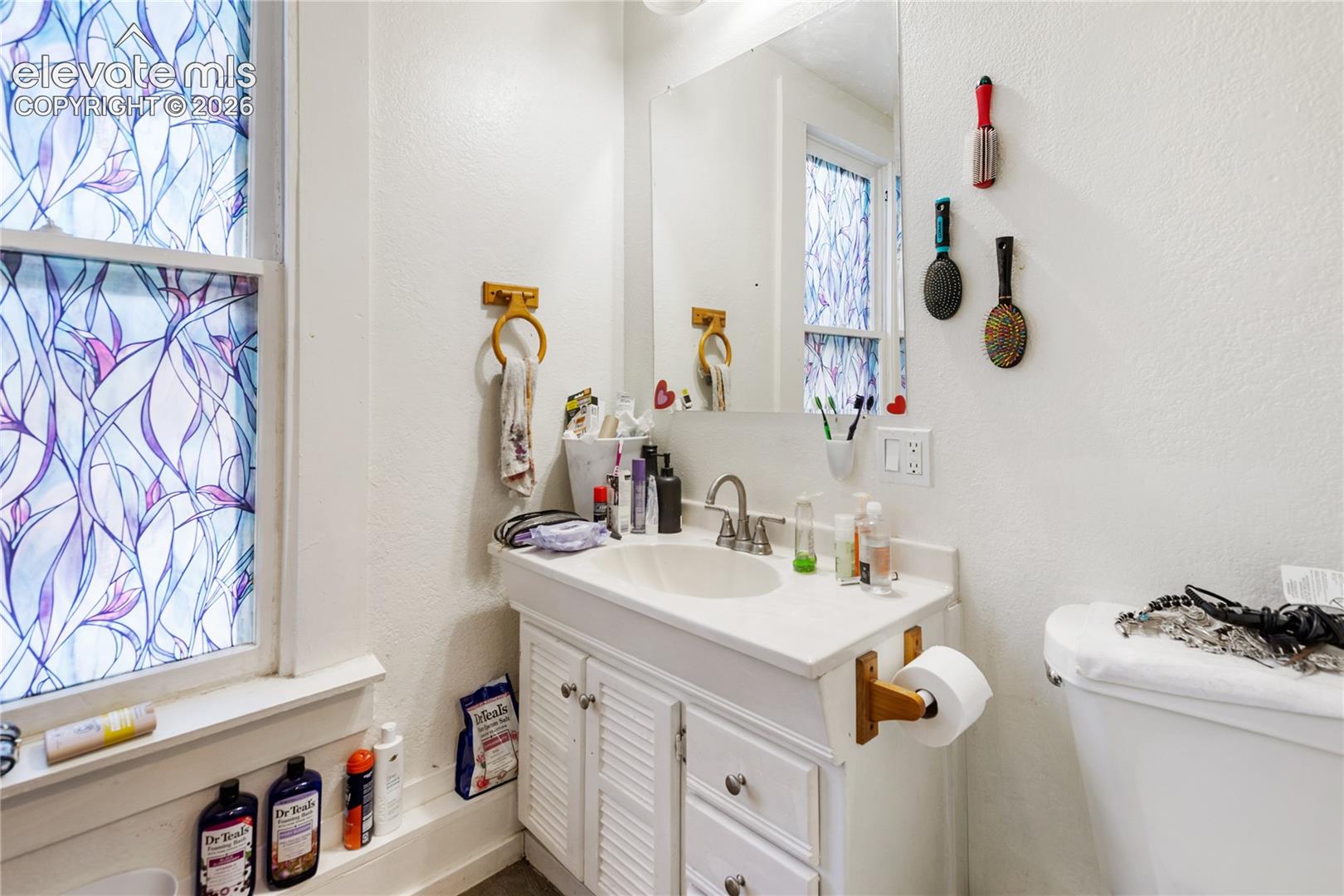 White vanity cabinet with louvered doors, integrated sink, wall-mounted mirror, and window with patterned privacy film