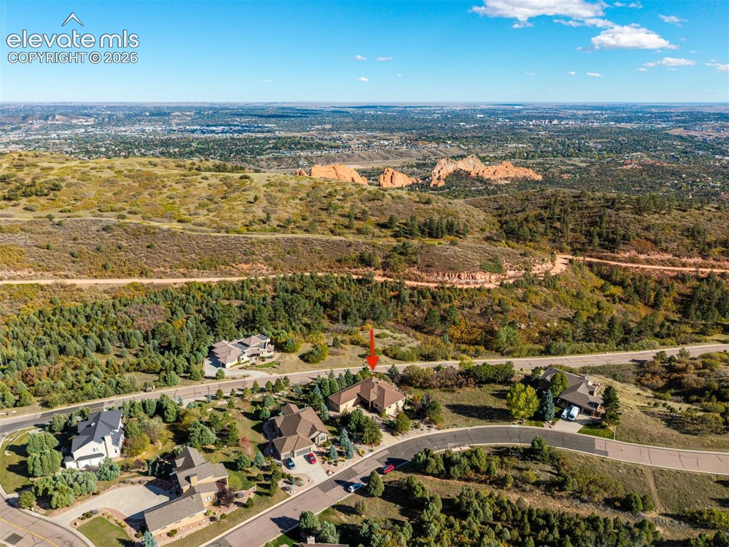 Aerial view highlighting Garden of the Gods, with its iconic red rock formations visible in the distance, and Rampart Range Road, a historic scenic mountain route winding through Pike National Forest toward Woodland Park. Situated on a .49-acre lot surrounded by mature trees and natural vegetation, the property features an expansive main-level balcony and covered walkout basement patio, offering multiple vantage points to enjoy the surrounding foothills and mountain scenery.