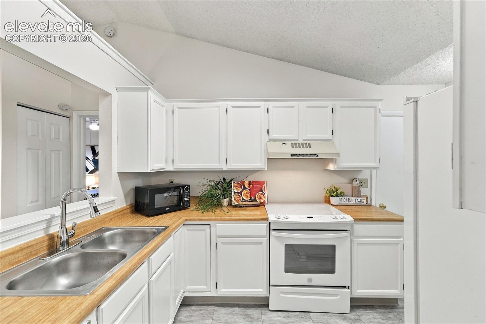Kitchen featuring white shaker-style cabinetry, wood-finish countertops, stainless steel double-basin sink with pull-down faucet, vaulted ceiling, and tile flooring