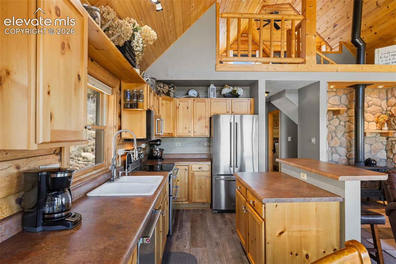 Kitchen featuring custom natural wood cabinetry, solid surface countertops, a dual-basin sink with a gooseneck faucet, stainless steel appliances, and wood-finish flooring, complemented by a stone-faced hearth and a loft area with log railings