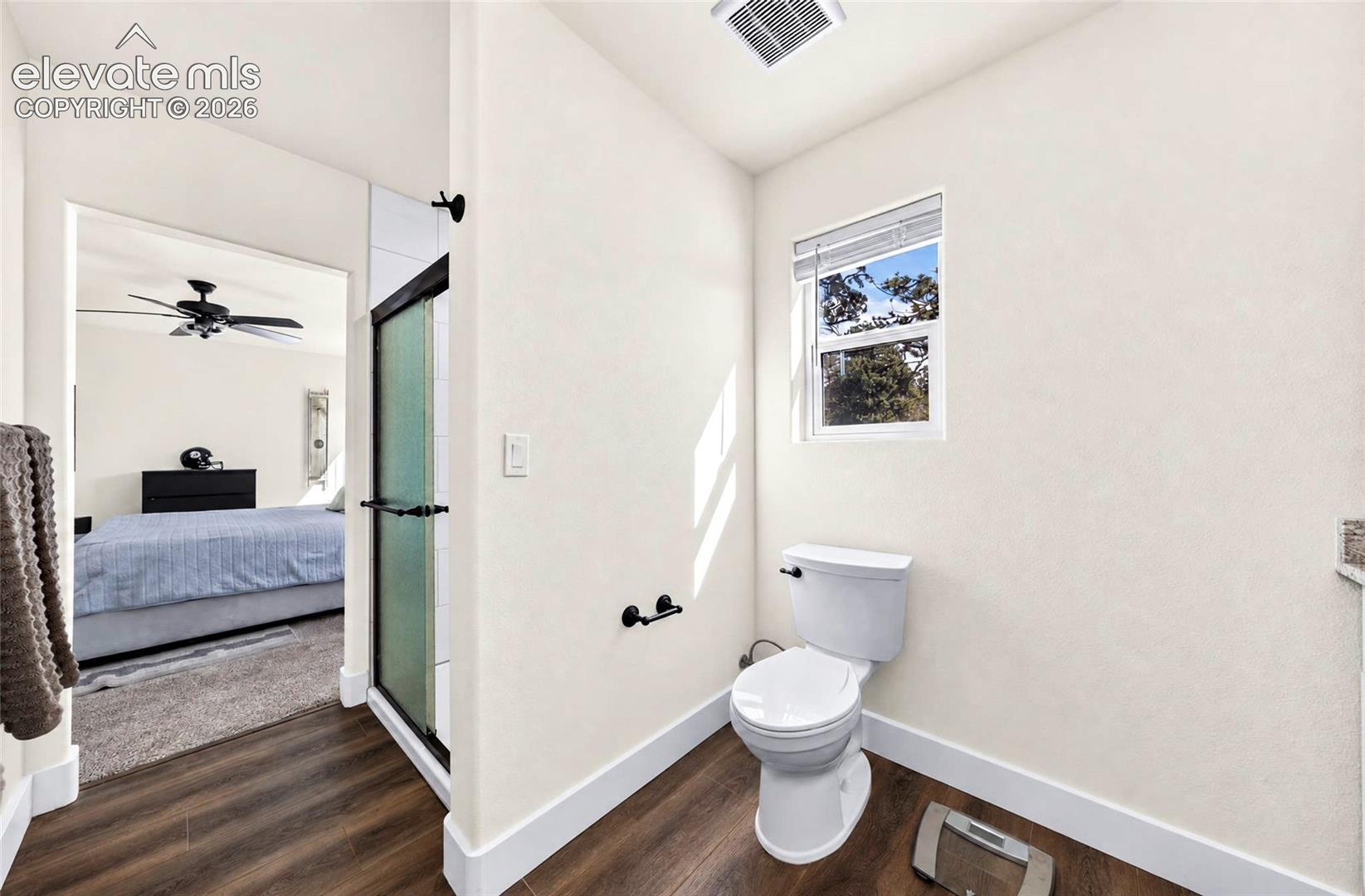 Bathroom featuring wood-finish flooring, a white porcelain toilet, a glass-enclosed shower with a matte black frame, and a single-hung window with horizontal blinds