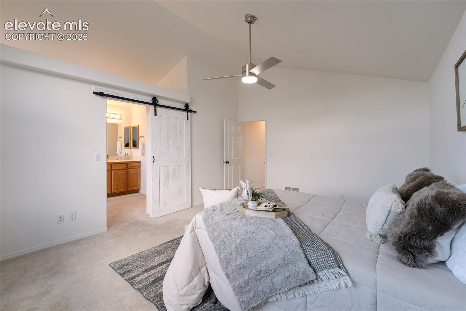 Vaulted ceiling, ceiling fan with light fixture, light-toned carpeting, white barn door, wood-finish vanity