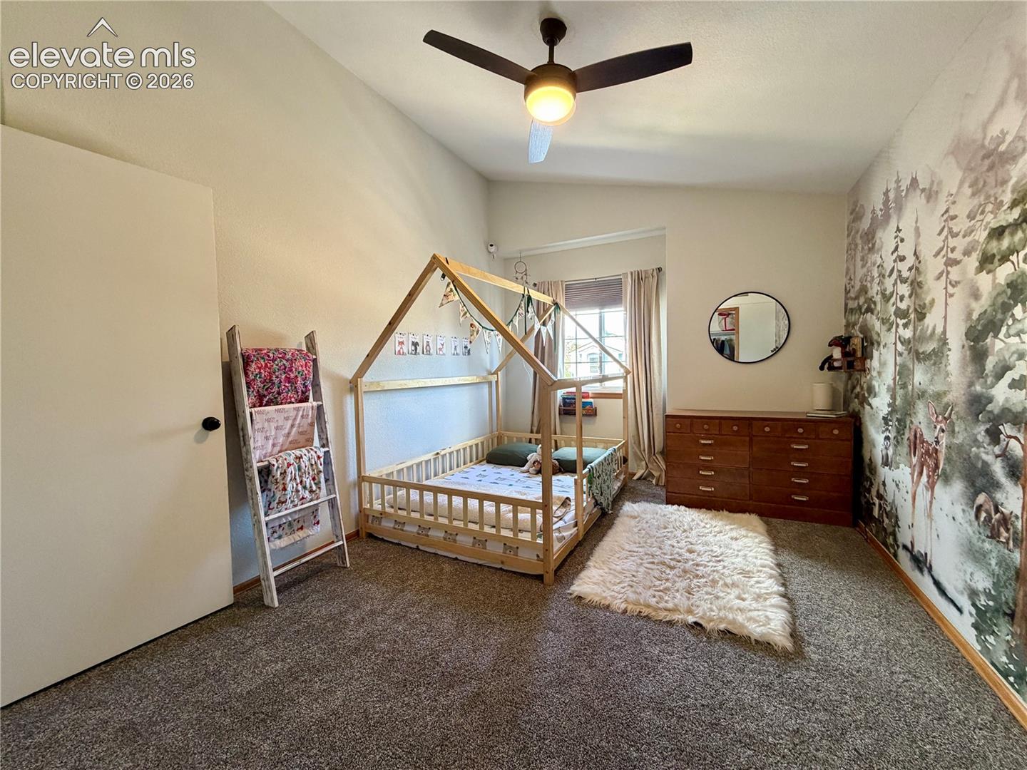 Carpeted bedroom featuring a ceiling fan with integrated lighting, a wall mural, and a window with natural light