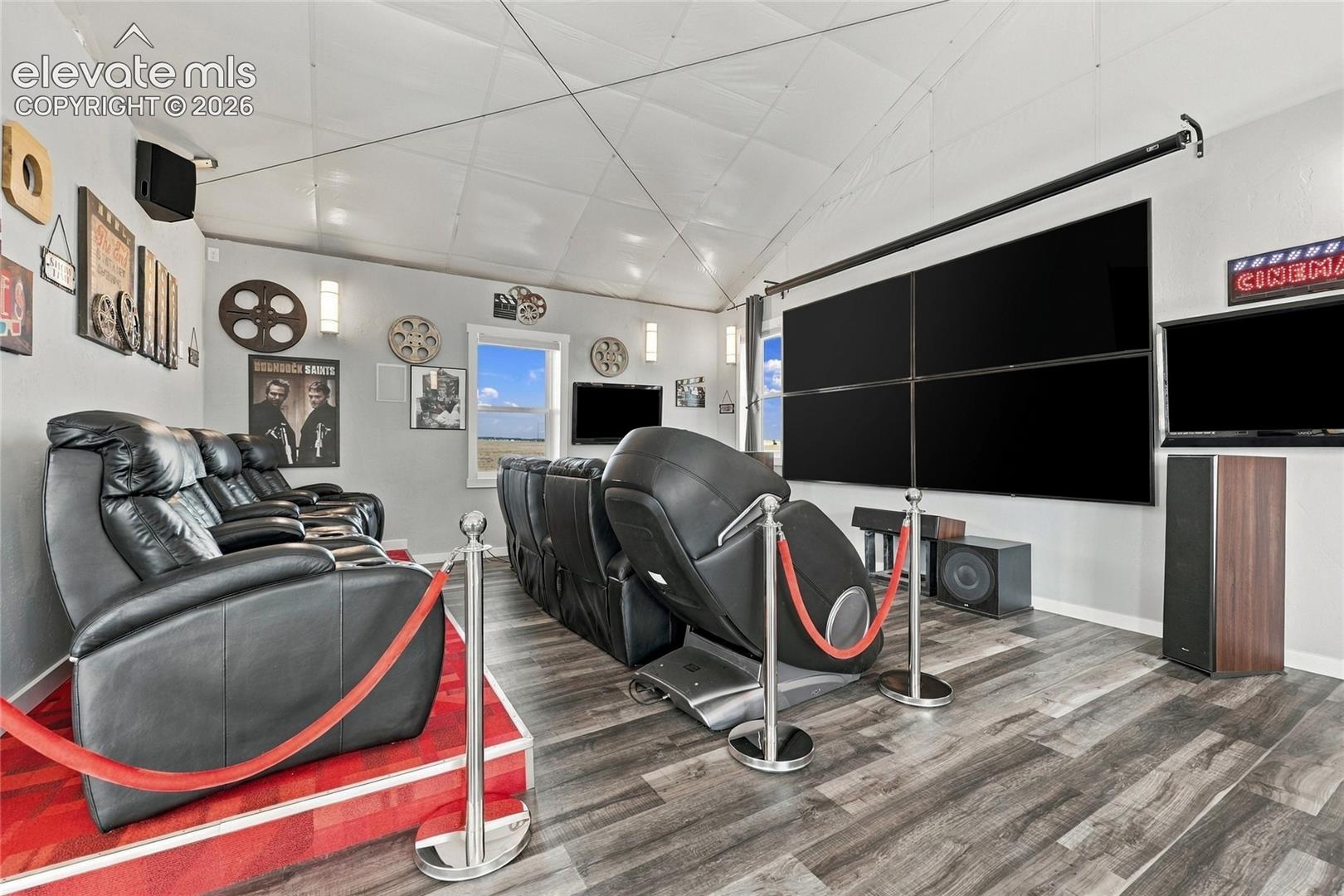 Media room featuring wood-finish flooring, vaulted ceiling, multi-panel wall-mounted screen display, and stepped platform staging