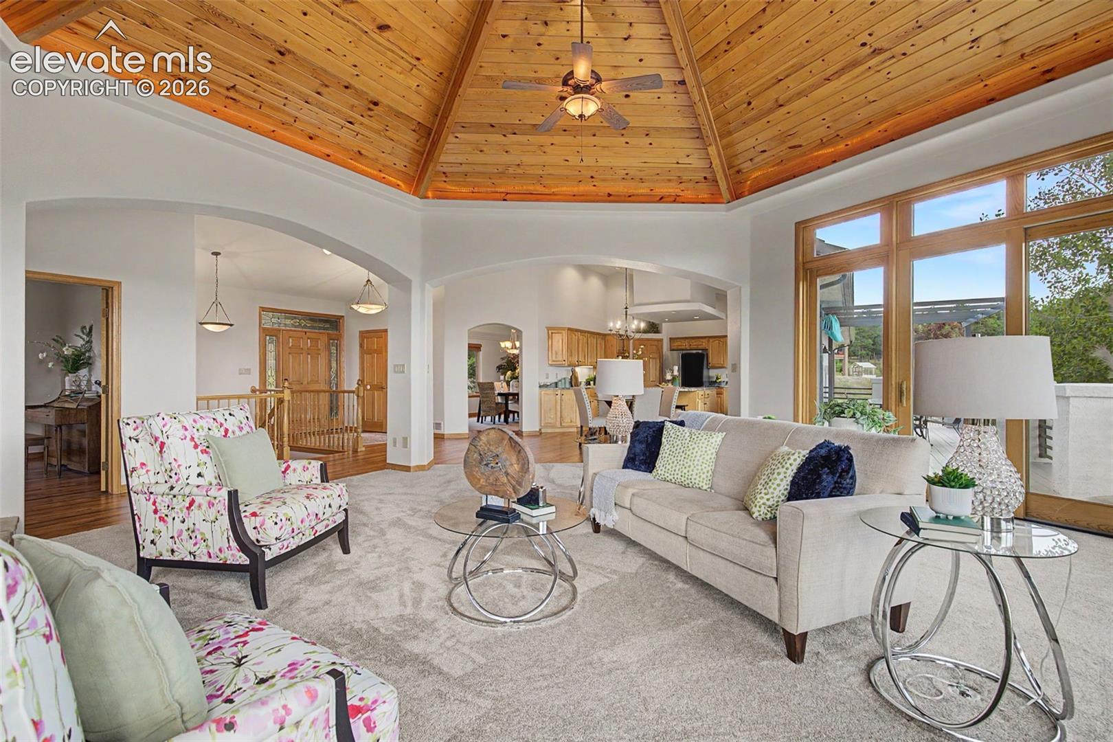 Expansive living area featuring a vaulted wood-paneled ceiling, large windows, arched doorways, light-toned carpet flooring, and a ceiling fan