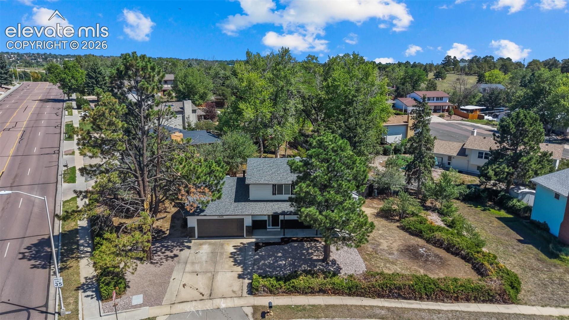 Aerial view of a contemporary home situated on a landscaped lot featuring mature trees, a paved driveway, and professionally designed hardscaping with stone and planting beds