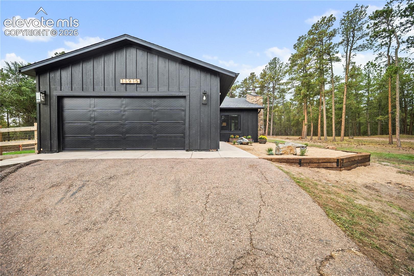 Black board-and-batten siding, asphalt driveway, concrete garage apron, and tiered timber landscape retaining wall