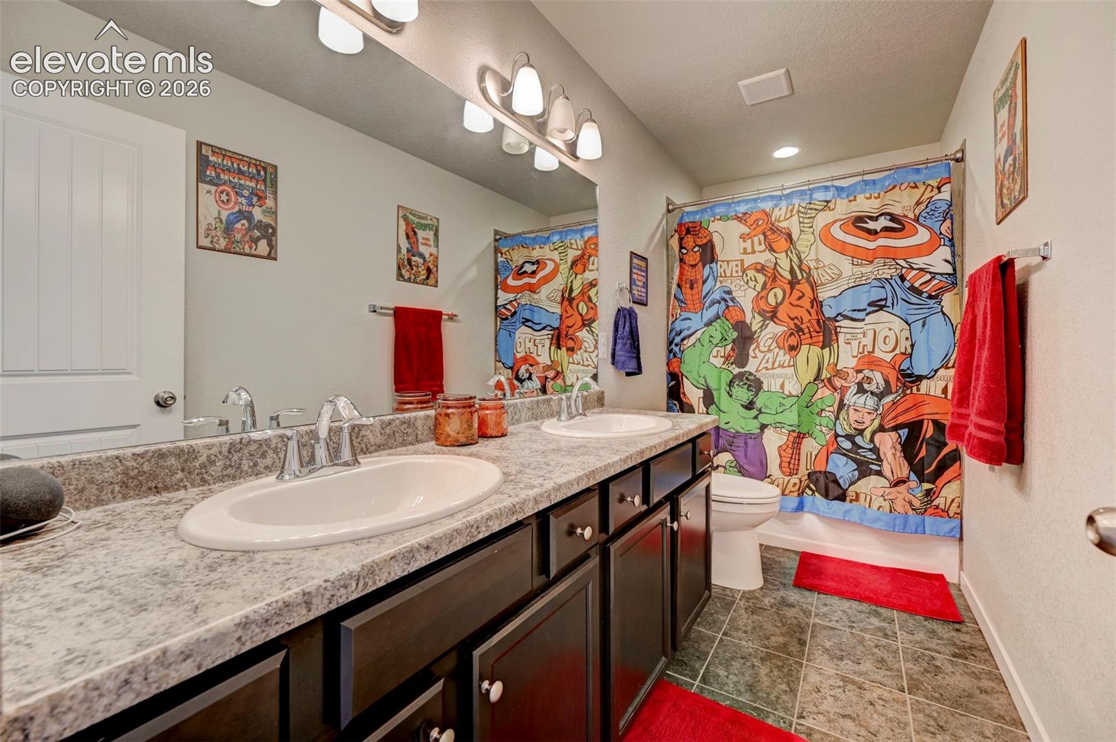Dual-sink vanity with stone-finish countertop, dark-toned cabinetry, full-width wall mirror, multi-light fixture, and tile flooring
