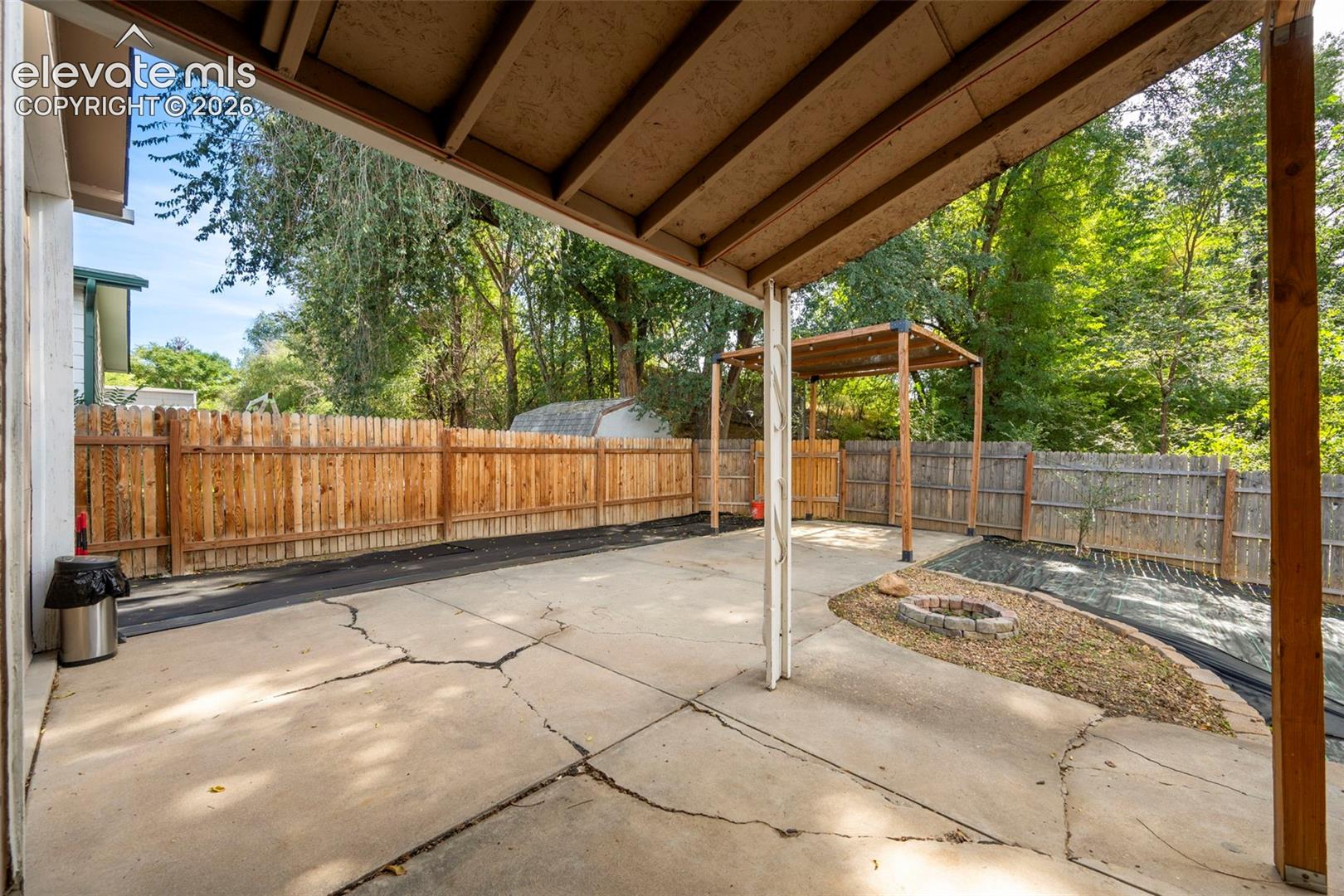 Covered concrete patio featuring a wooden pergola, wood fencing, and a stone-lined fire pit area