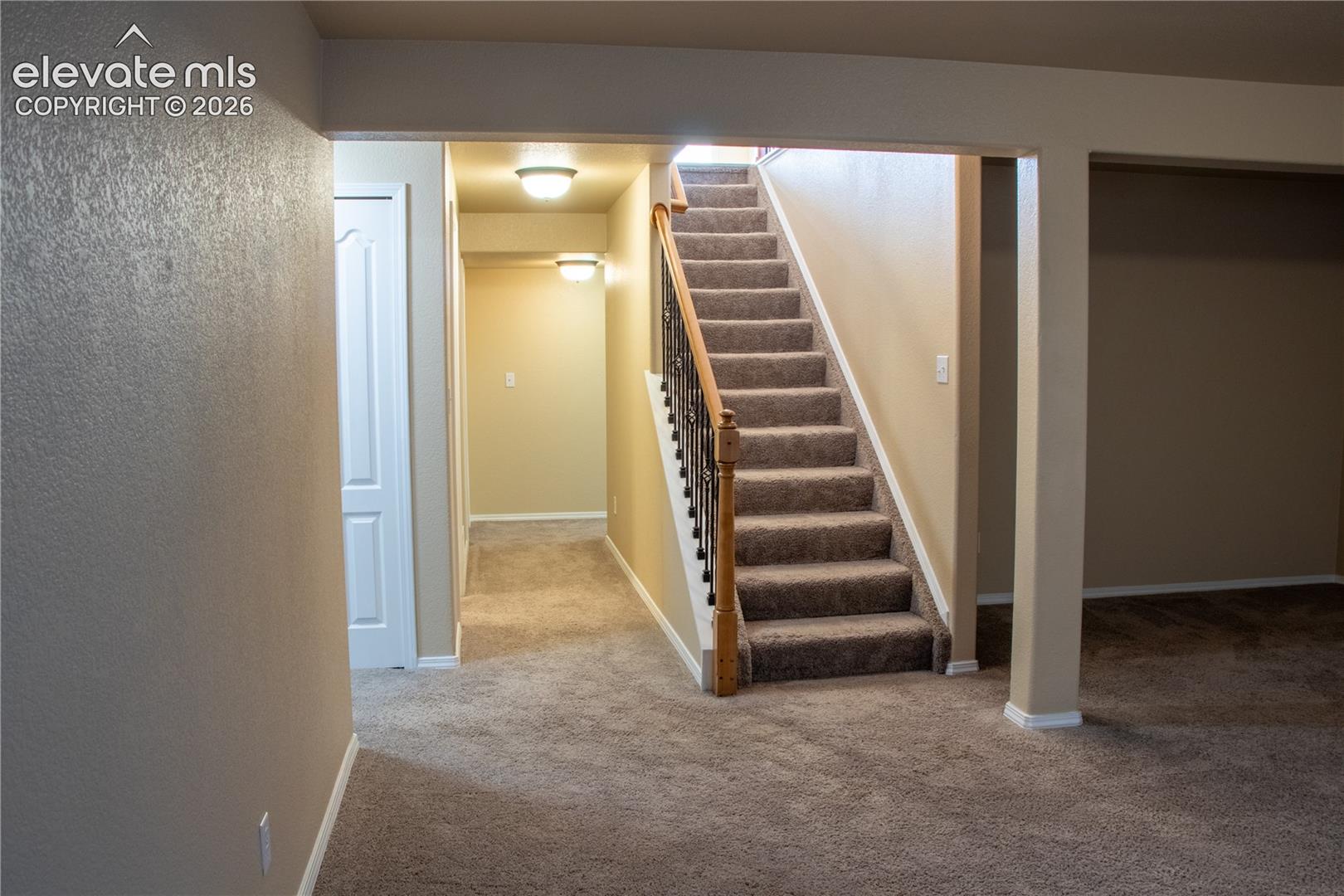 Carpeted staircase with wood handrail and wrought iron balusters, complemented by recessed lighting and a carpeted hallway