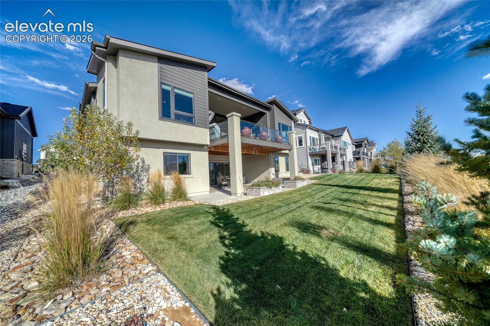 Multi-story exterior featuring a second-level balcony with glass panel railing, stucco and horizontal siding, stone landscape accents, and an open grass area