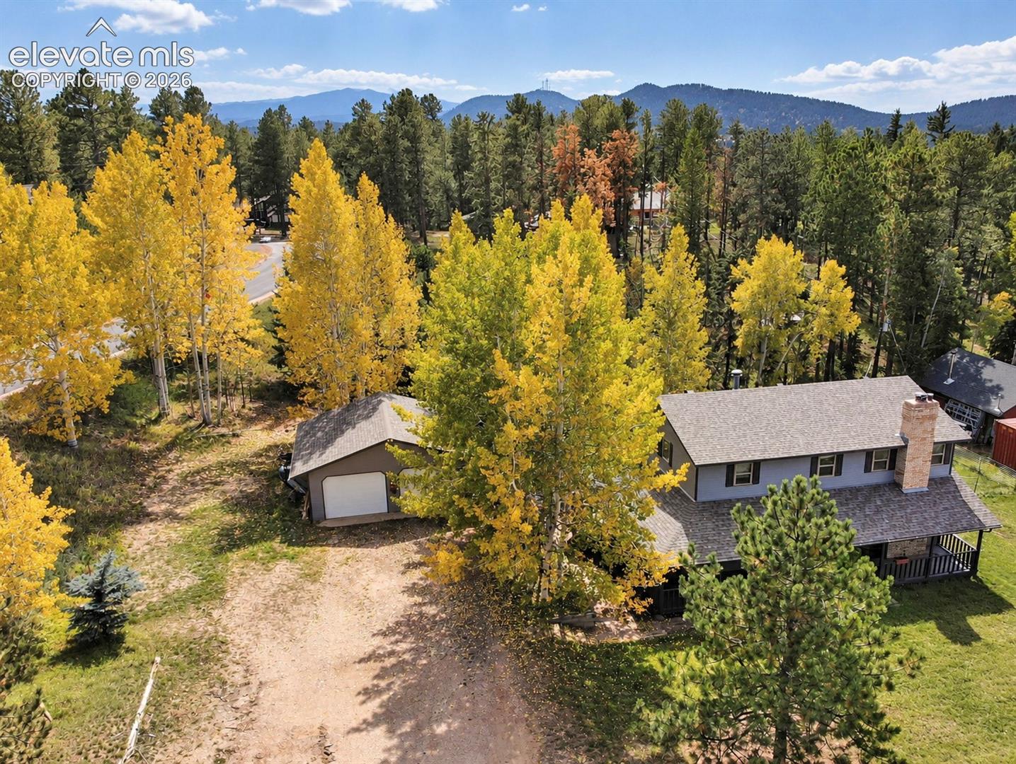Two-story gray siding residence featuring a prominent brick chimney, an attached covered porch, a separate garage with a white overhead door, mature trees, and mountain scenery