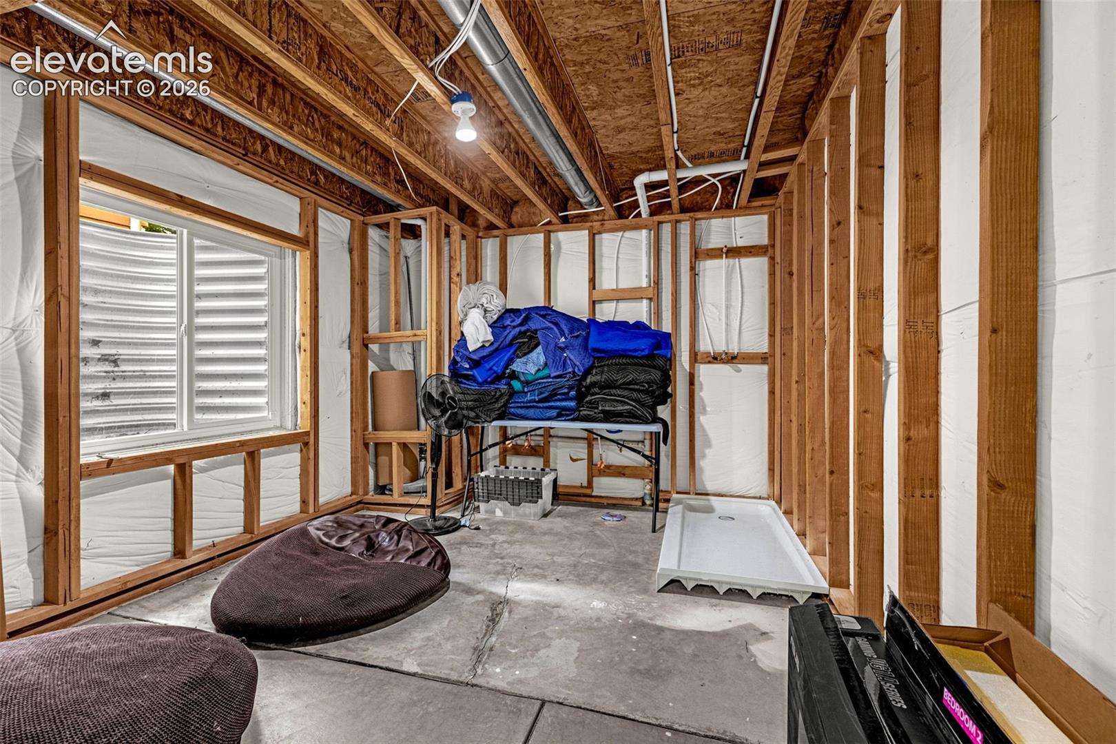 Unfinished space featuring exposed timber framing, concrete flooring, an egress window with a corrugated steel well, and a pre-installed shower pan