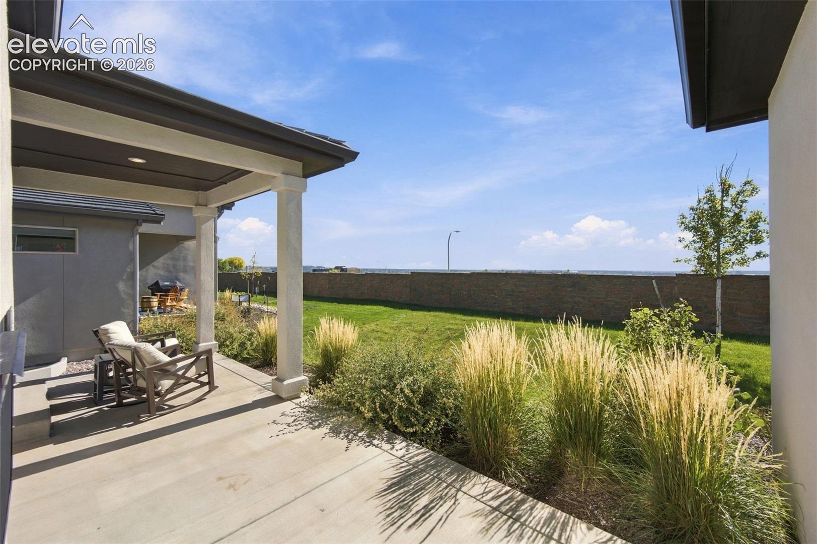 Covered concrete patio featuring recessed lighting and square columns, overlooking a fenced backyard with landscaped grasses and a lawn