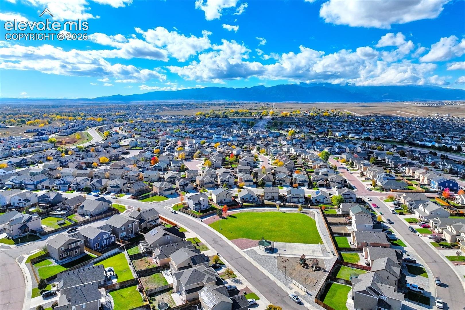 Aerial perspective of residential neighborhood featuring a central community park with manicured lawn, paved pathways, and mountain scenery