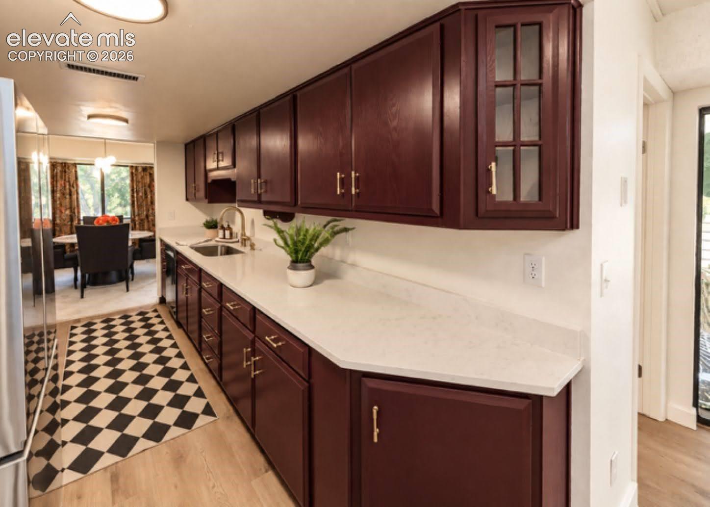 Kitchen featuring deep-toned cabinetry, light solid-surface countertops, metallic hardware, an upper cabinet with glass-panel doors, and wood-finish flooring