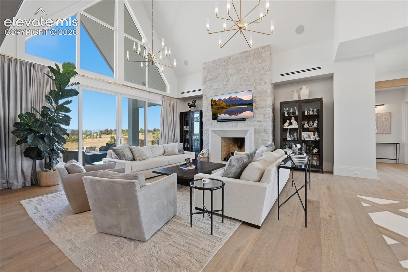Vaulted ceilings with expansive floor-to-ceiling windows, stone-clad fireplace, wood-finish flooring, and multi-arm chandelier lighting