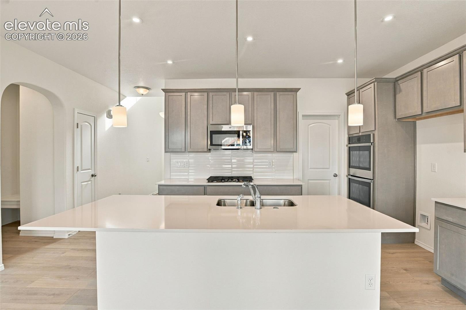 Kitchen featuring a central island with a solid surface countertop, light-toned shaker-style cabinetry, a white tile backsplash, stainless steel built-in double wall ovens, and wood-finish flooring