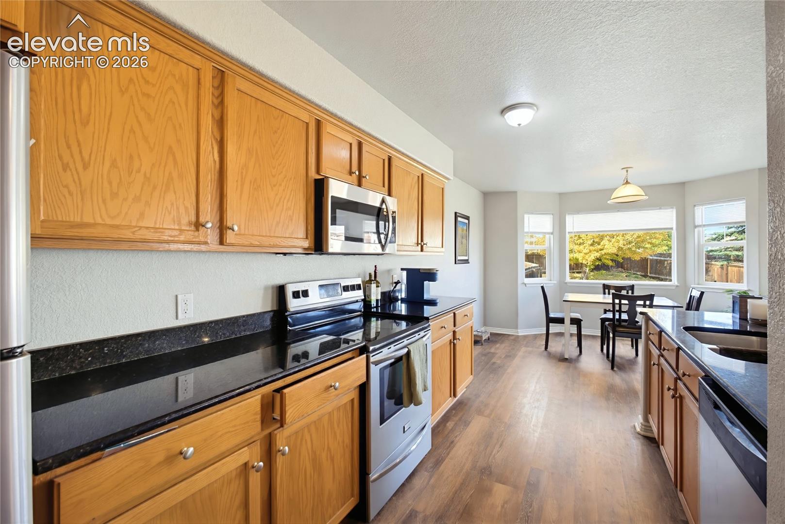 Kitchen featuring wood cabinetry, dark stone-finish countertops, stainless steel appliances, wood-finish flooring, and a bay window