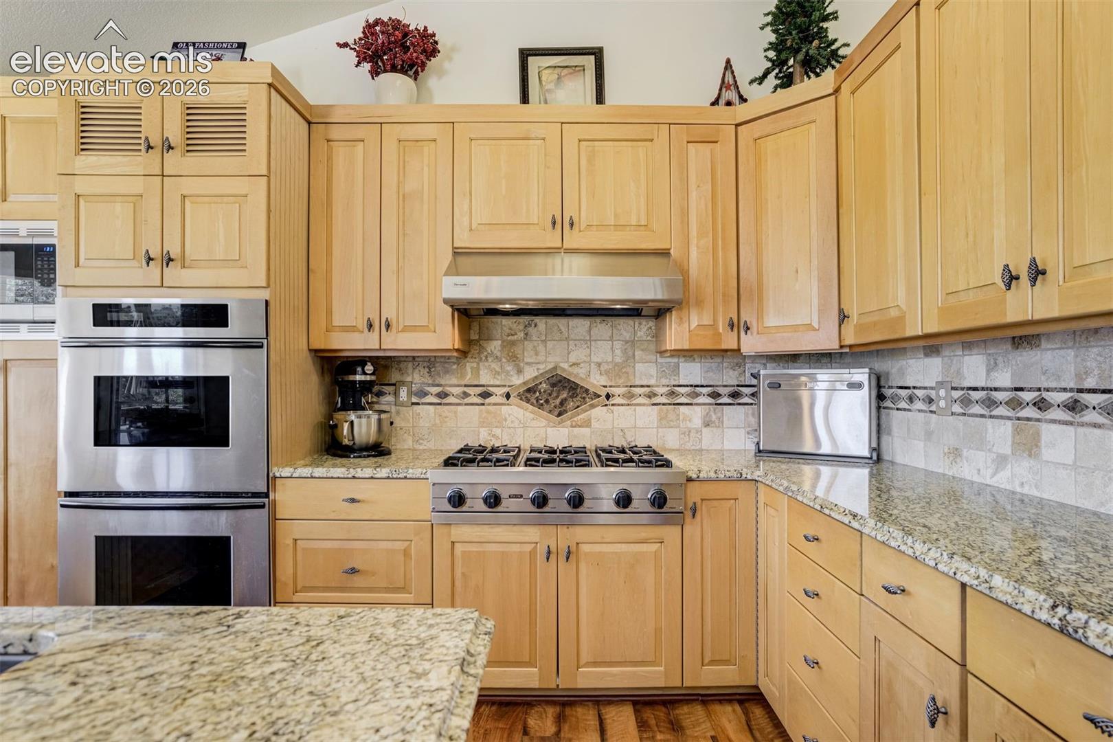 Light wood cabinetry featuring shaker-style doors and upper louvered accents, natural stone countertops, stainless steel double wall oven, stainless steel gas cooktop with matching range hood, and a decorative tile backsplash with stone inlay