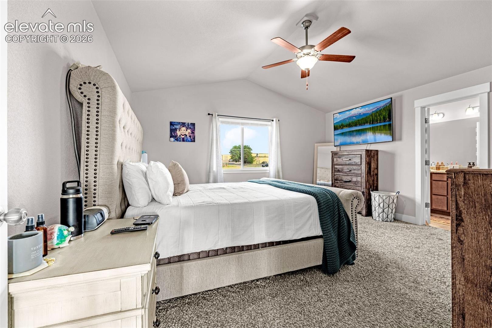 Carpeted bedroom featuring a vaulted ceiling with a ceiling fan, a window, and an open doorway revealing a bathroom with a dual vanity
