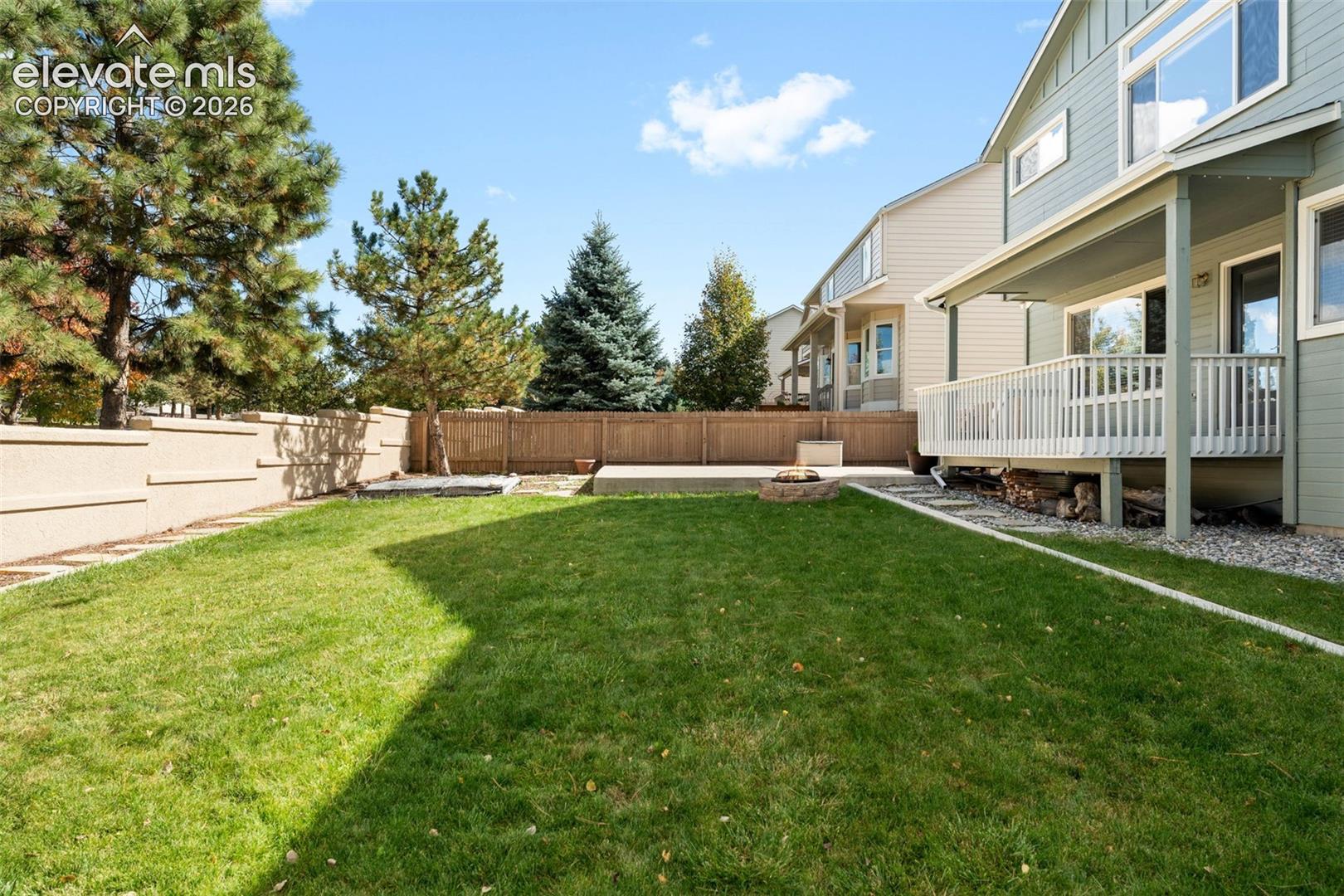 Backyard featuring an elevated wooden deck with railing, a stone-lined fire pit, concrete patio area, perimeter fencing, and mature pine trees