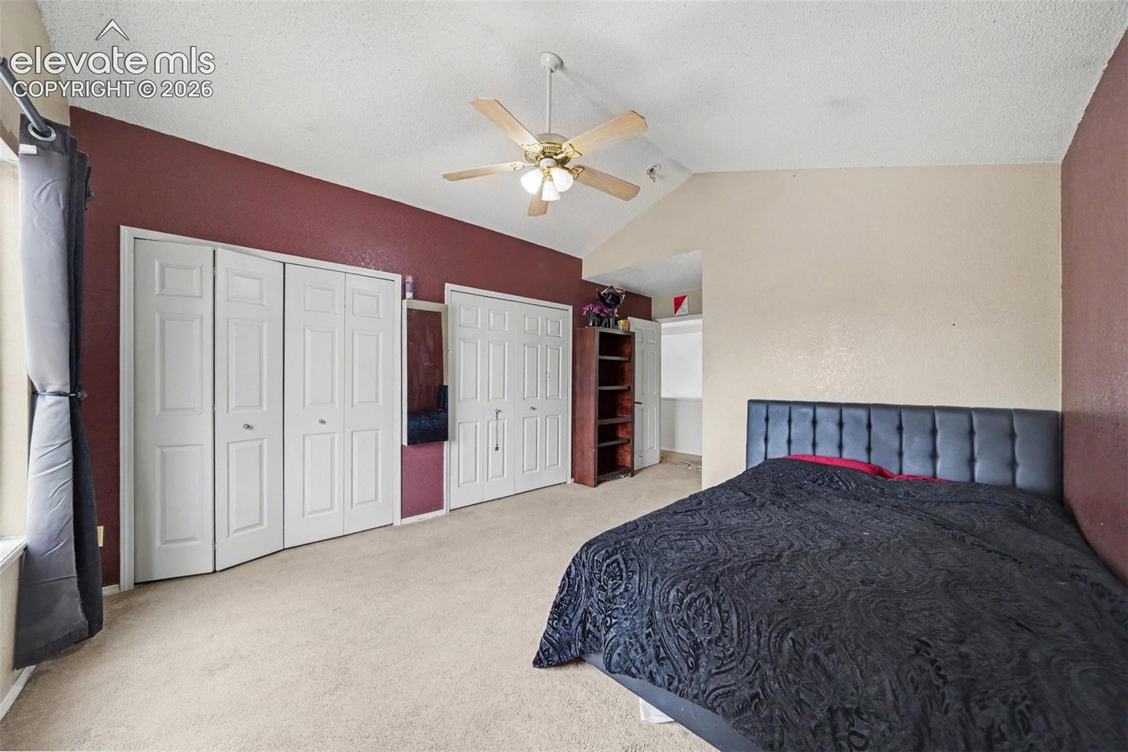 Vaulted ceiling featuring a central multi-blade ceiling fan, expansive wall-to-wall closet storage with white paneled bifold doors, and neutral carpeting