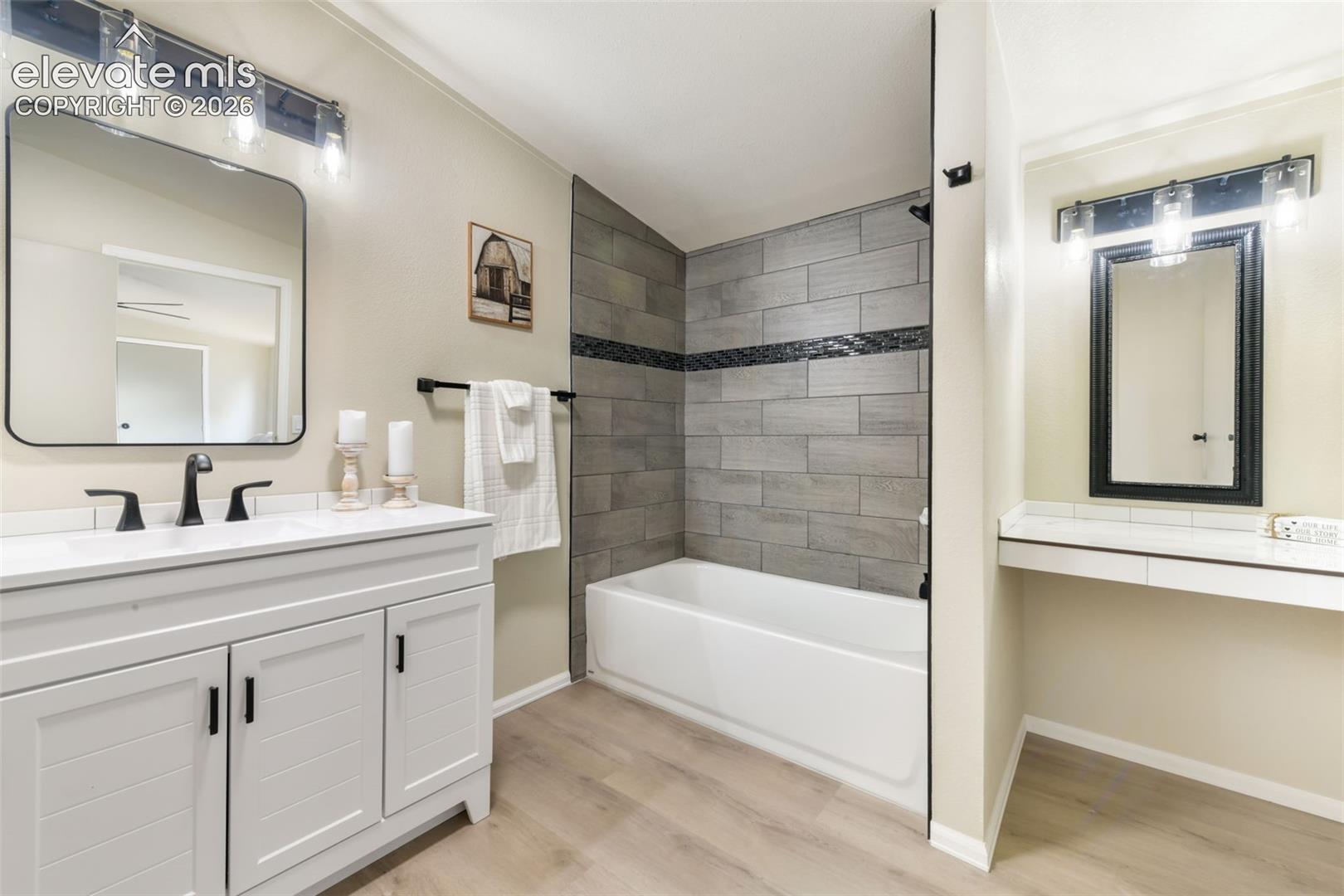 Wood-finish flooring, white vanity with black hardware, rectangular mirror with black frame, matte black faucet, and a tiled shower with an accent band