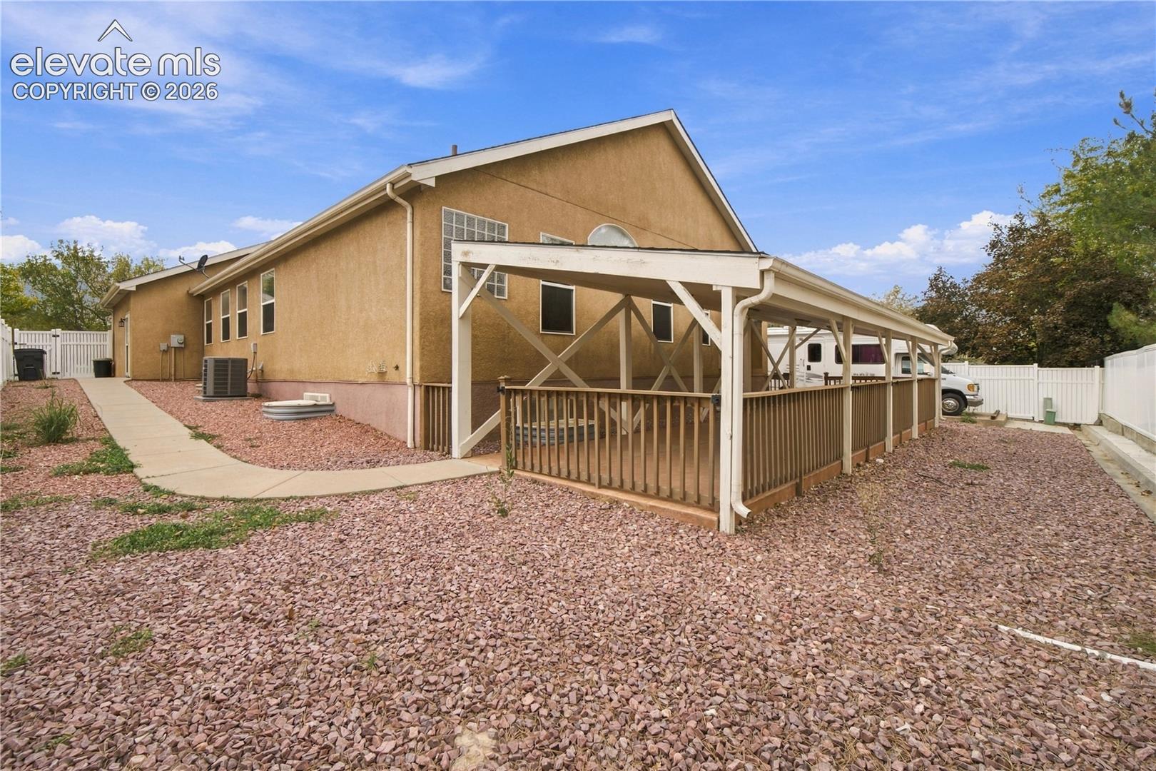 Stucco exterior featuring an expansive covered wood deck, gravel landscaping, concrete walkway, and white vinyl privacy fencing