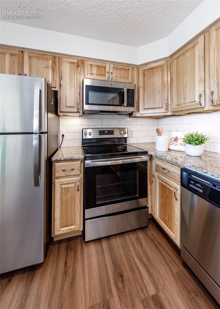 Kitchen featuring wood cabinetry, stone countertops, white subway tile backsplash, wood-finish flooring, and stainless steel appliances