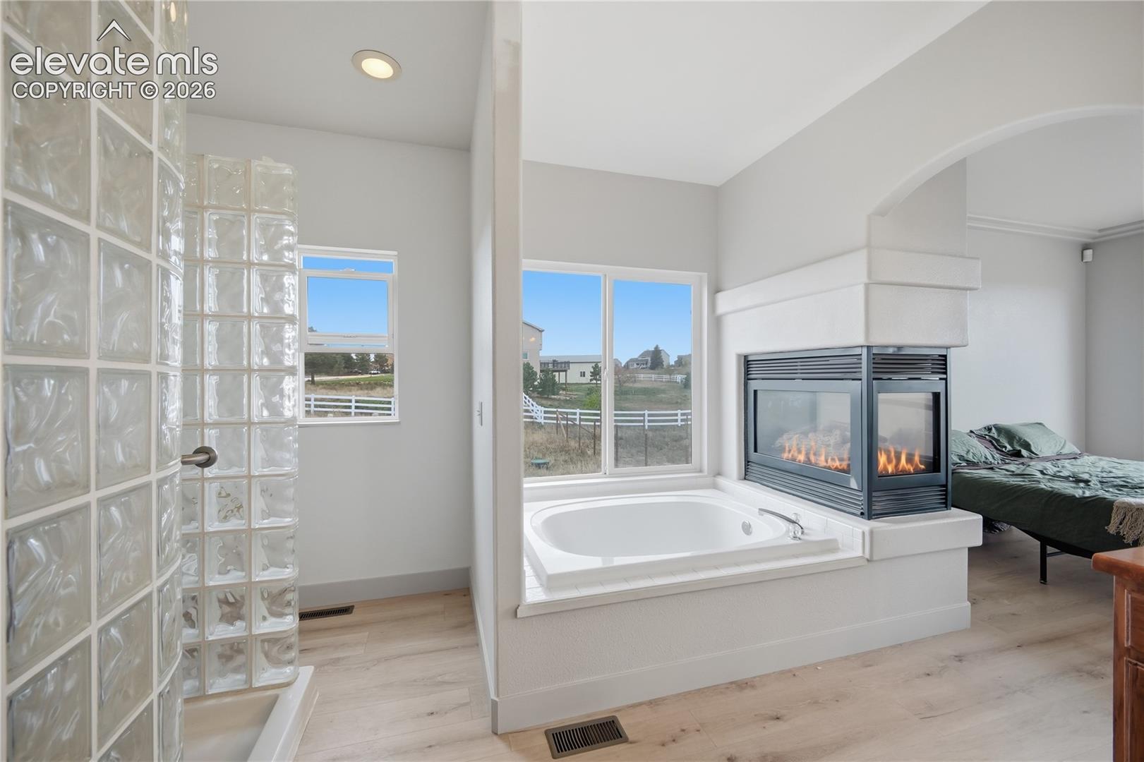 Deep soaking tub featuring a tiled surround, dual-sided fireplace, curved glass block wall, and wood-finish flooring