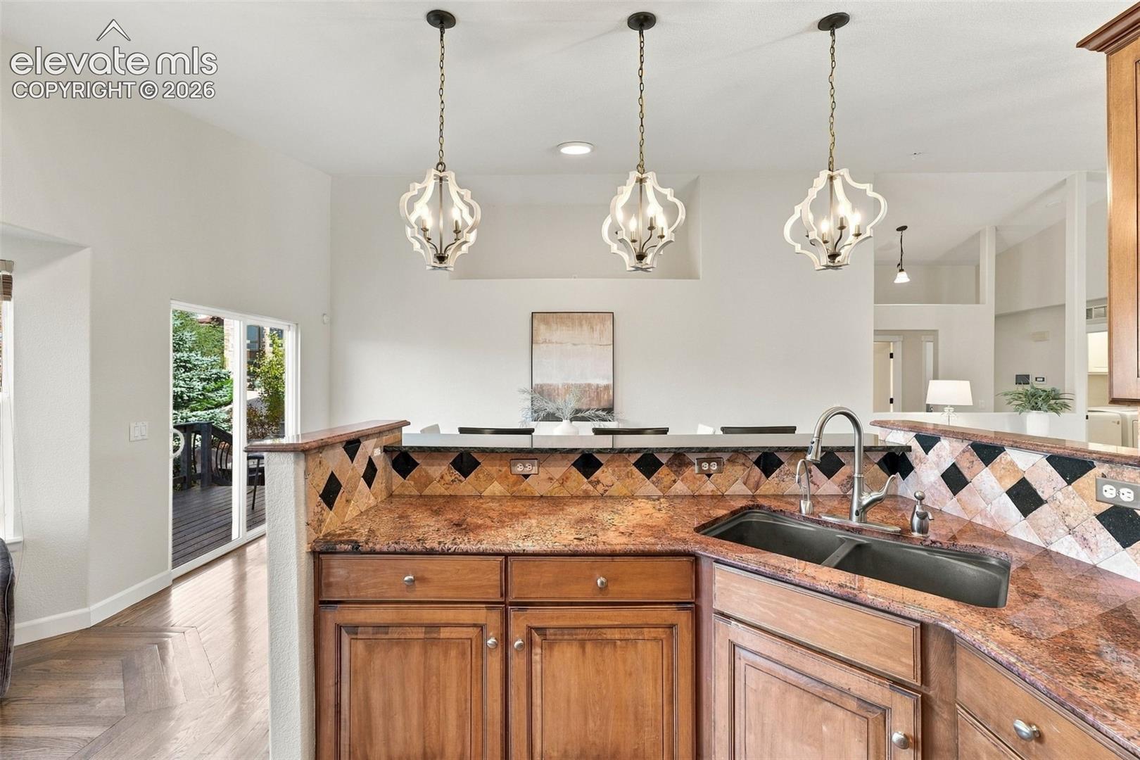 Kitchen featuring wood cabinetry, stone countertops, diamond-patterned tile backsplash, undermount dual-basin sink with pull-down faucet, and trio of decorative pendant lights