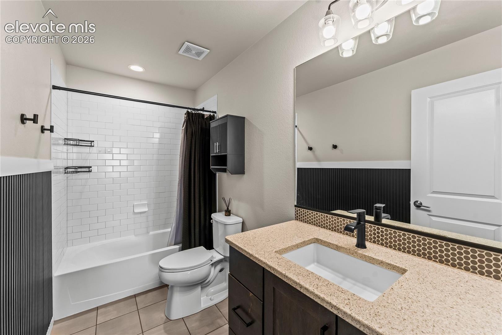 Bathroom featuring a vanity with a rectangular sink, stone-composite countertop, and matte black faucet, accompanied by a tub with white subway tile surround, tile flooring, and dark beadboard wainscoting