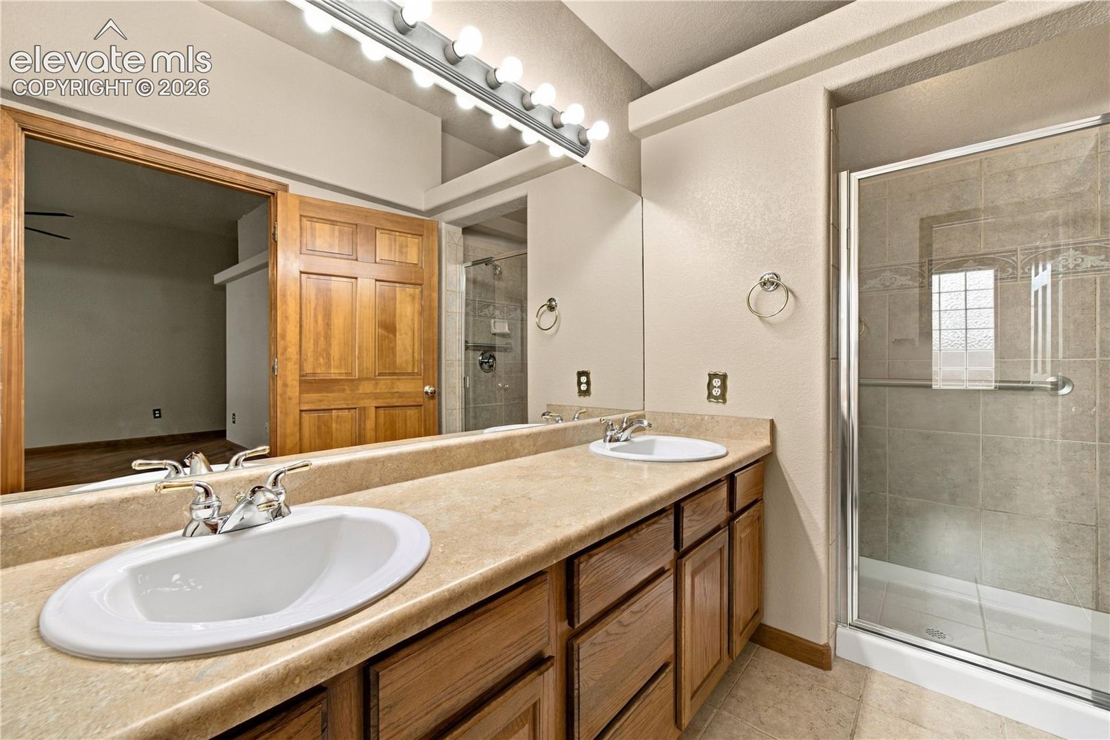 Dual-sink vanity with a solid-surface countertop, wood cabinetry, wall-to-wall mirror, and a walk-in shower with tiled surround