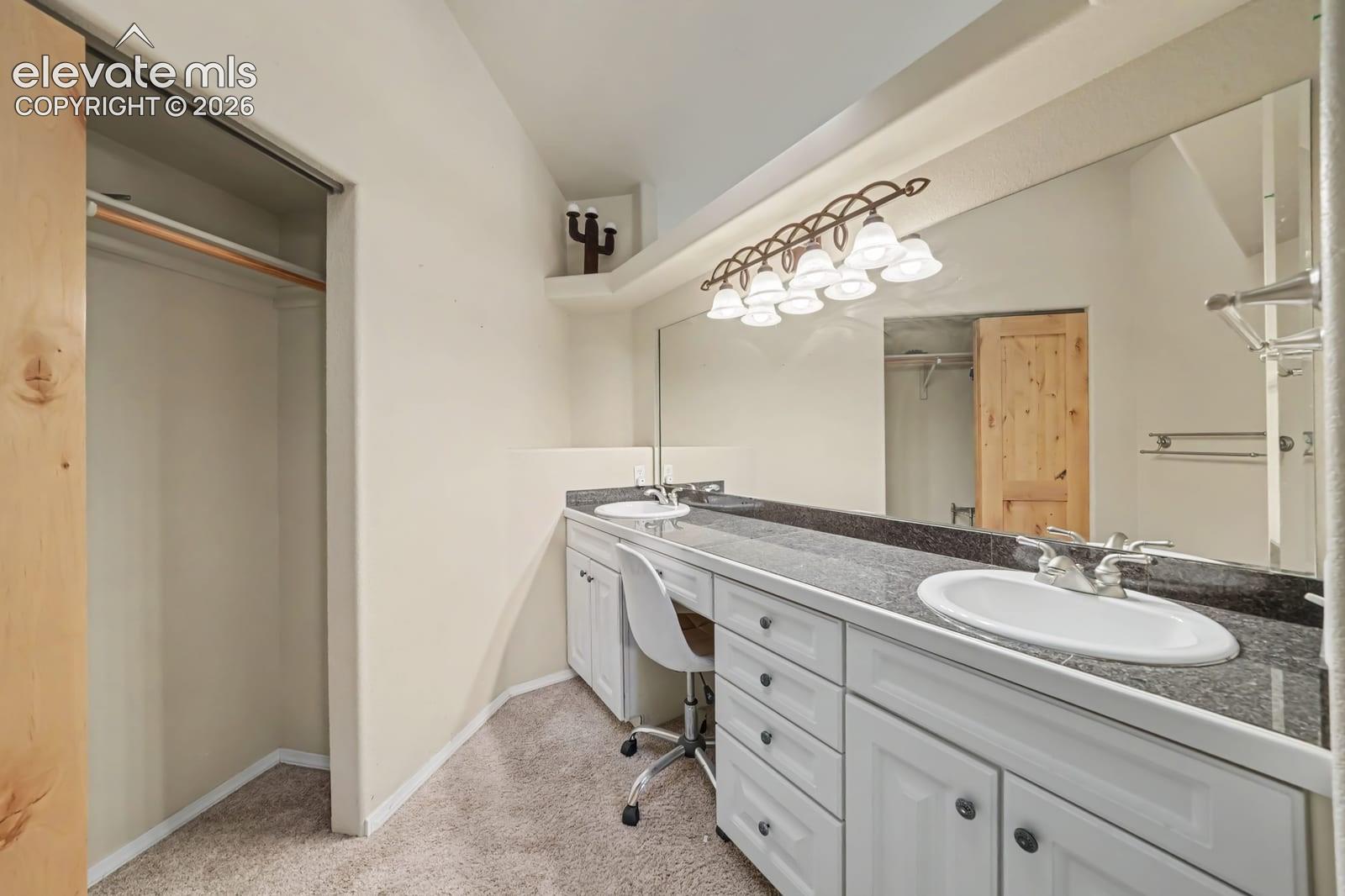 Extended vanity with white cabinetry, stone-finish countertops, dual integrated sinks, full-width mirror, and a recessed closet with built-in shelving